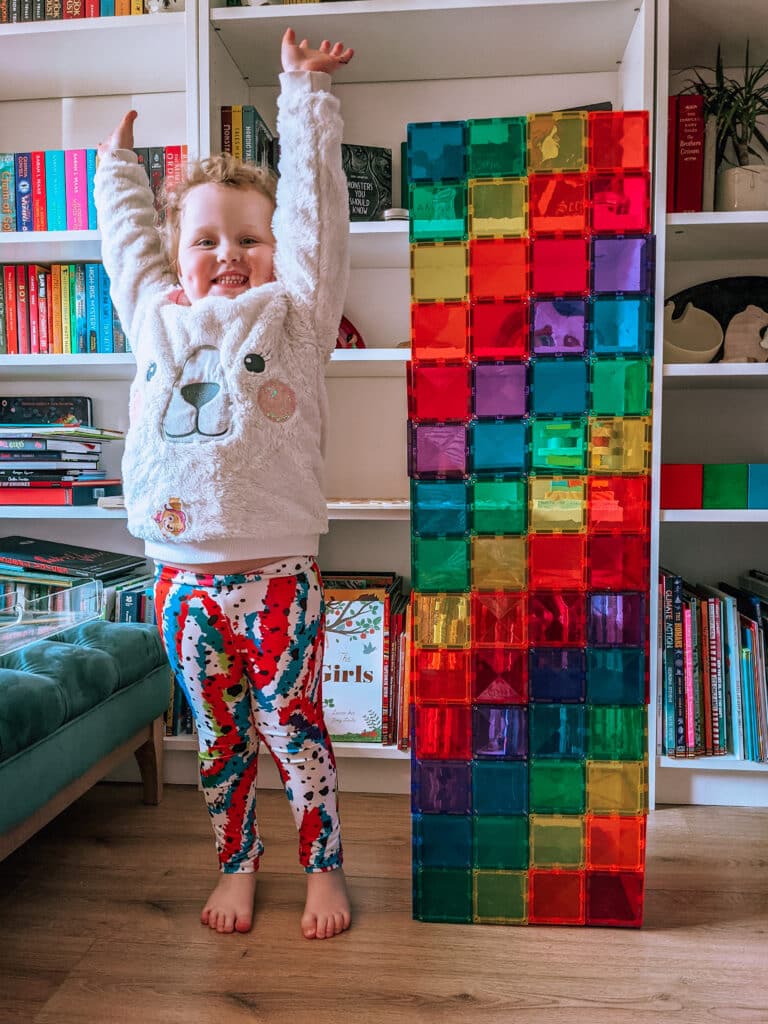 A child standing with hands in the air next to a Rainbow Connetix Tower made of standard Connetix Squares that is taller than her.