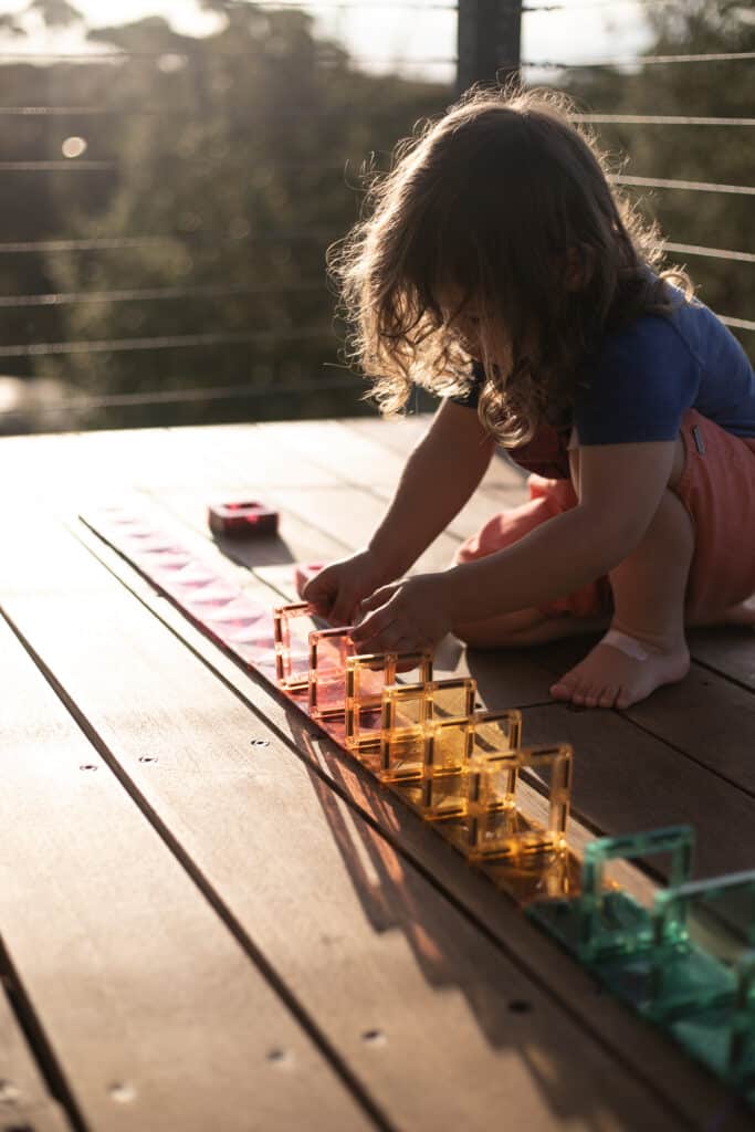 child lining up Connetix pastel square tiles in a domino track outdoors on a deck.